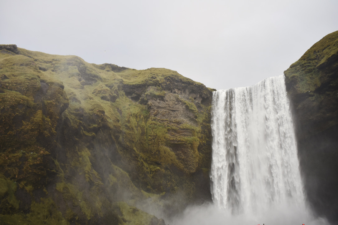 A photo of Skógafoss, a waterfall in Iceland. The waterfall is wide, set between two moss-covered rocks. There is a lot of spray in the air and the weather is overcast.