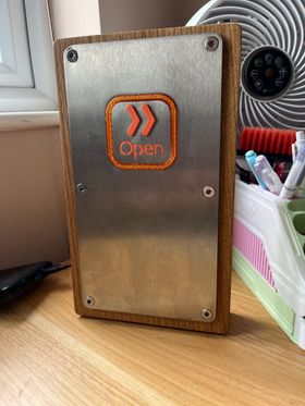 A wooden box containing a metal panel with a distinctive-looking square button in the middle. The button has an orange ring around it, and the word 'Open' with a pair of chevrons.