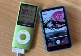 A green iPod nano Gen 5, plugged in with its wide 20-pin connector, playing The Lottery from Knives Don't Have Your Back by Emily Haines. On its right, a small blue and black Hiby R1 Mp3 player, shorter but chunkier. It's got a higher-res screen, and is playing Glass Ceiling by Metric from the album Live It Out.