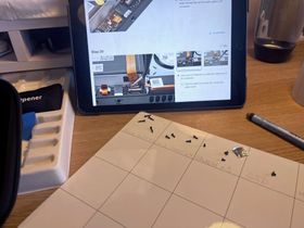 An ipad is propped up on the desk showing instructions for repairing the Steam Deck. In front is a white project mat with a grid on, and various tiny screws in the top 4 squares with what they are for written underneath: Back outer x4, Back inner x4, shield x3, SSD