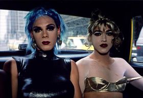 Two drag queens sit in the back of a taxi. One of them is wearing a tight metallic tank sleeveless top and has a short, choppy blue wig; she is wearing large silver heart-shaped earrings, thick dark eyeshadow and deep crimson lipstick. The second drag queen is wearing a long-sleeved ripped white fishnet crop top under a metallic bra, with a curly blonde updo wig, very pale face powder with dark eyeshadow and dark red lips. The straps of her bra are hanging off her shoulders.