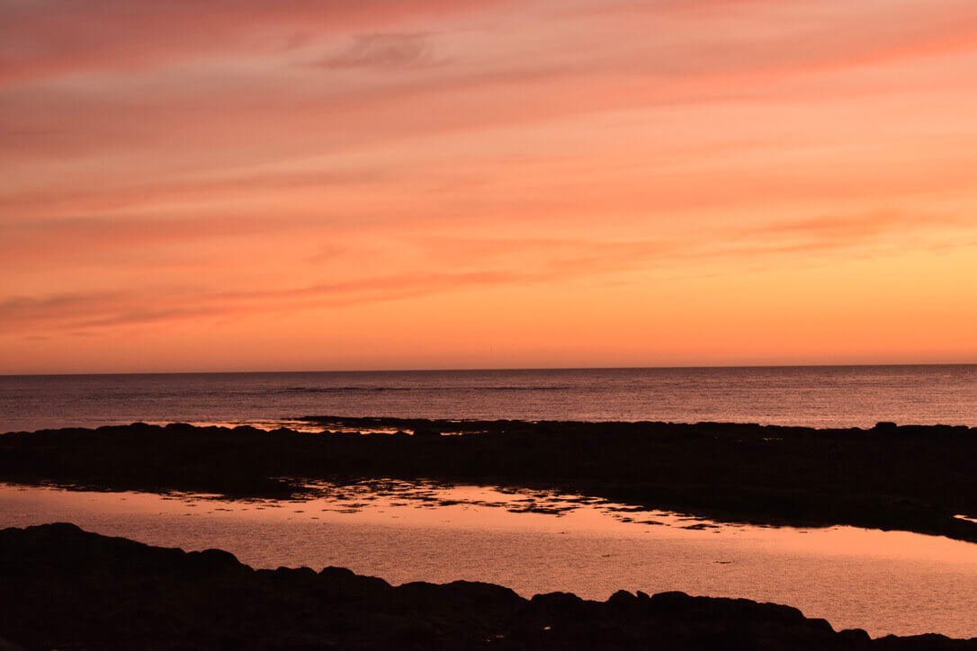 A bright orange sunset, looking out onto the water with some very dark stretches of beach at the bottom, so dark they are black.