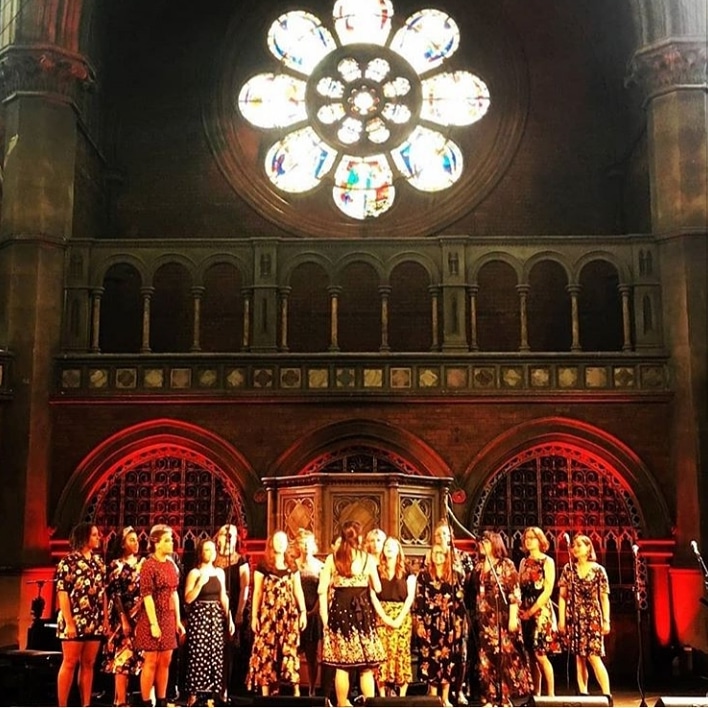 a photo of the choir on a stage at the Union Chapel in Islington, with the iconic round flower shaped window above them