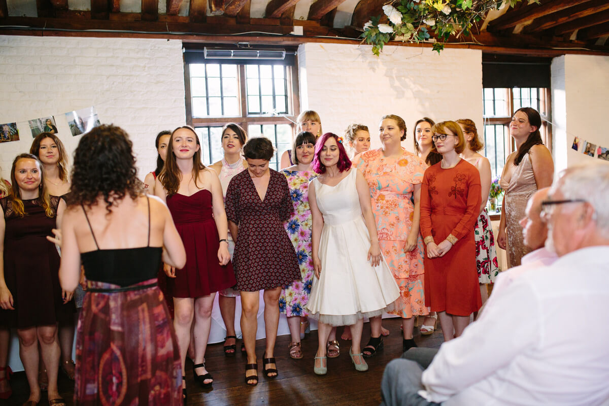 Sophie in a knee length white wedding dress, surrounded by women in smart dresses, peparing to sing. they stand together in a converted barn, the exposed rafters visible above them.