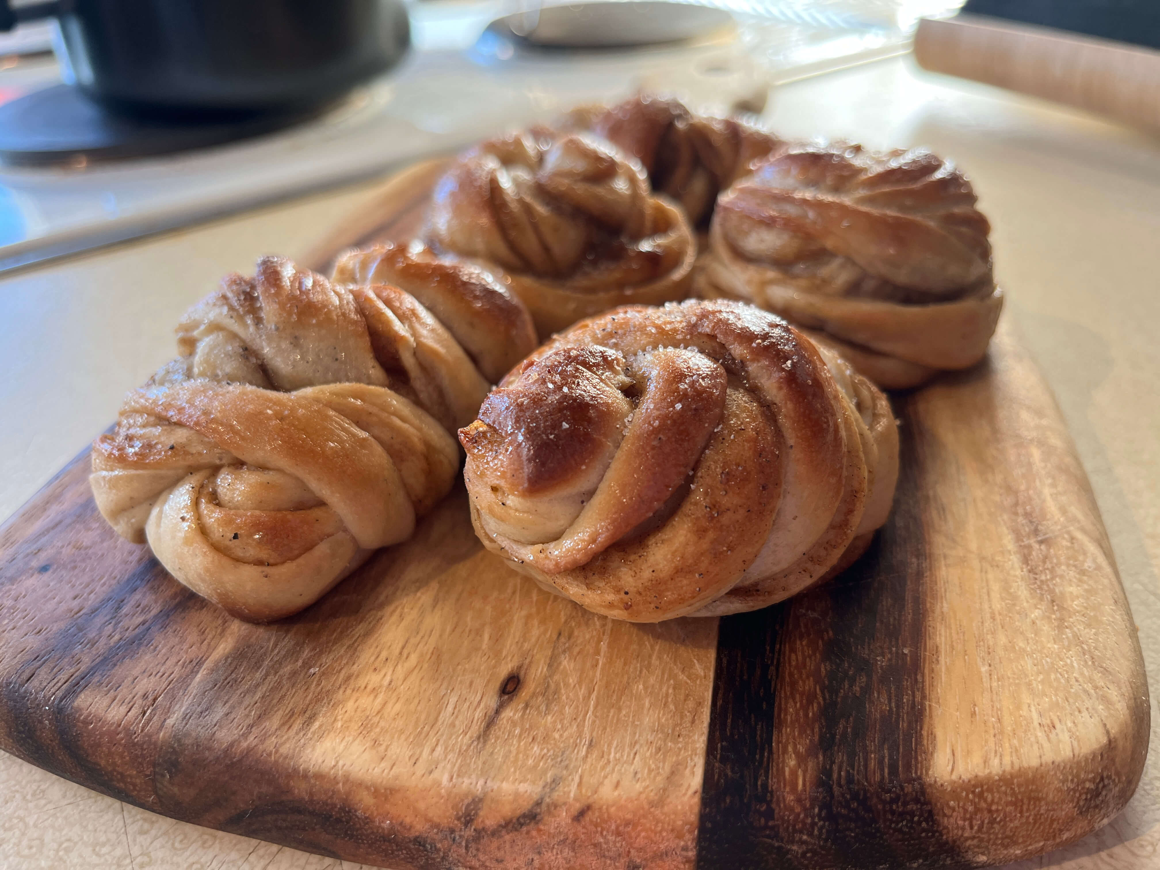 Small knotted cardamom buns sit carefully posed on a wooden board. They're glazed, and dusted with sugar.