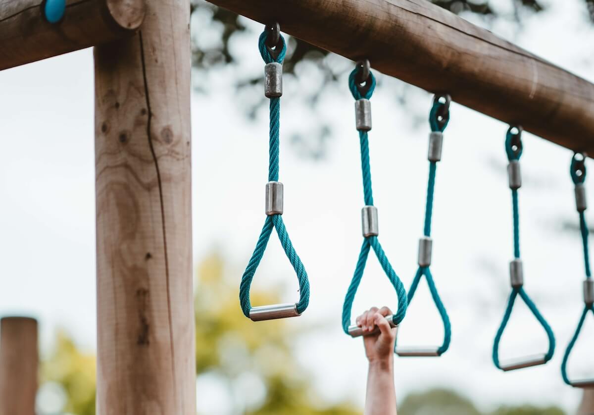 A child's hand clutches one of several handles hanging from a horizontal wooden pole on a jungle gym