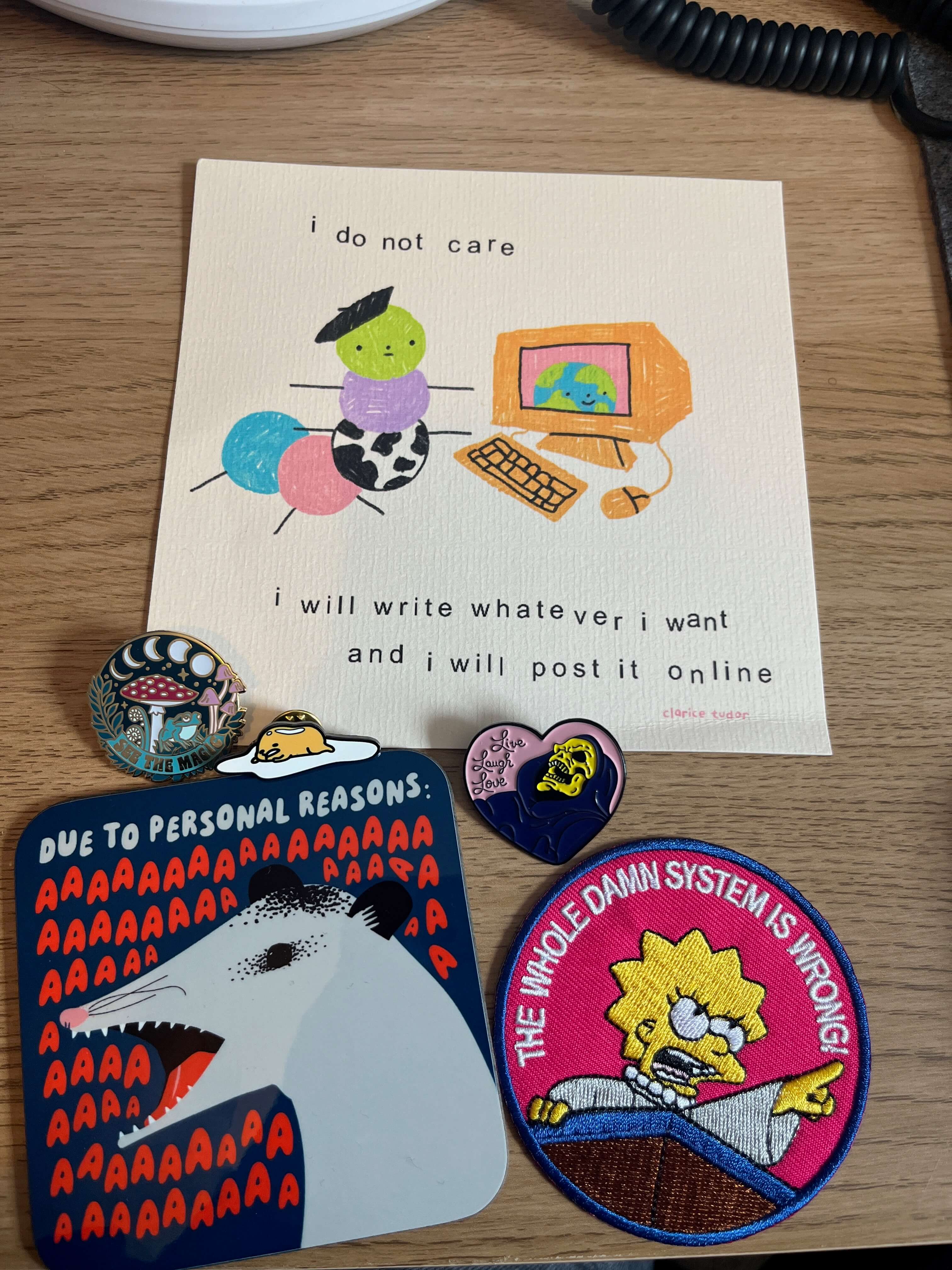 A collection of new purchases on a desk: a coaster featuring an opossum that says 'Due to personal reasons aaaaaaaaaaaaaaa'; an iron-on patch featuring Lisa Simpson pointing and saying 'The whole damn system is wrong!'; a pin badge of Gudetama being lazy and lying down; a heart-shaped pin badge featuring Skeletor that says 'Live Laugh Love'; a pin badge with a toadstool, a frog, and phases of the moon that says 'see the magic'; and a square print of a caterpillar wearing a beret in front of a computer with the caption 'I do not care, I will write whatever I want and post it online'.