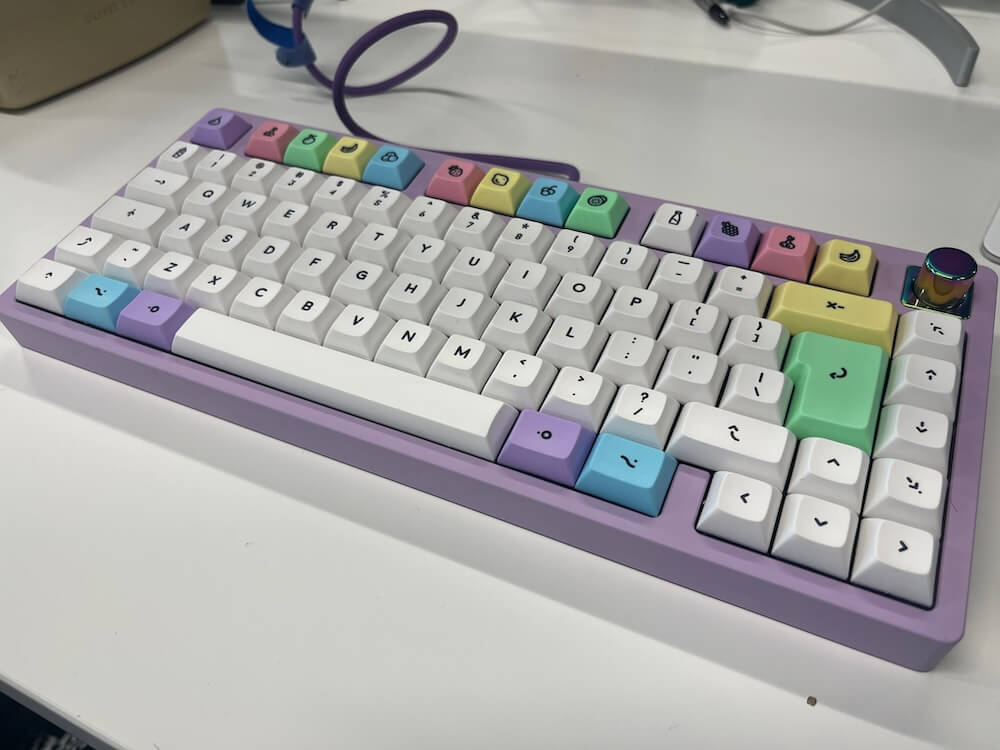 The keyboard on a white desk. It has a pastel purple case, and white keys with pastel coloured accents: a purple CMD key, a blue alt key, and a green ISO enter key. The F-key row are all pastel coloured fruits. On the top right is an iridescent purple knob.