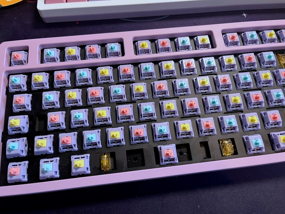 The keyboard with no keycaps on, showing the switches underneath. They have pastel purple housing with pastel pink, yellow and blue stems