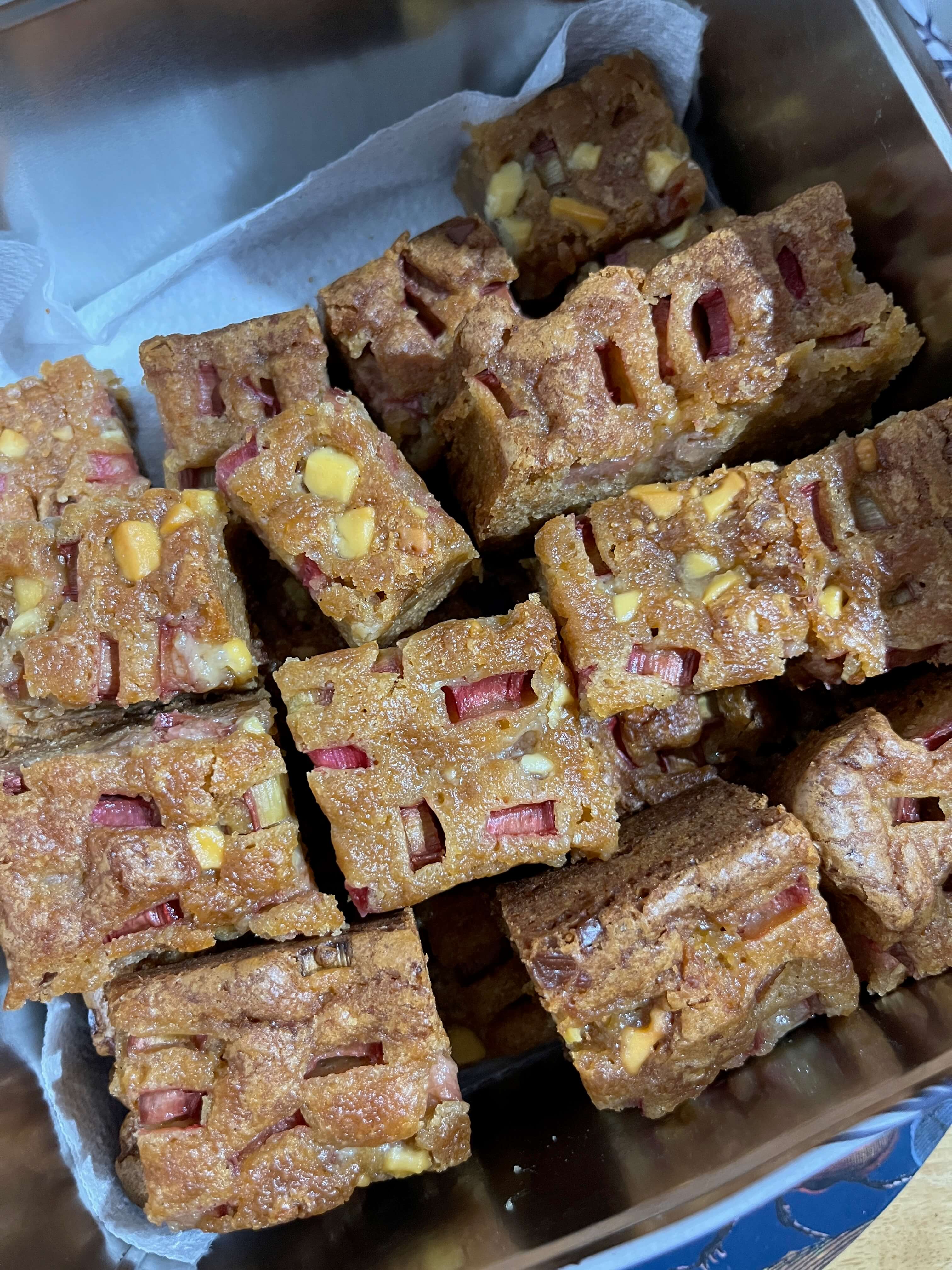 A tin of rhubarb and custard blondies, golden coloured squares studded with white chocolate and slices of rhubarb.