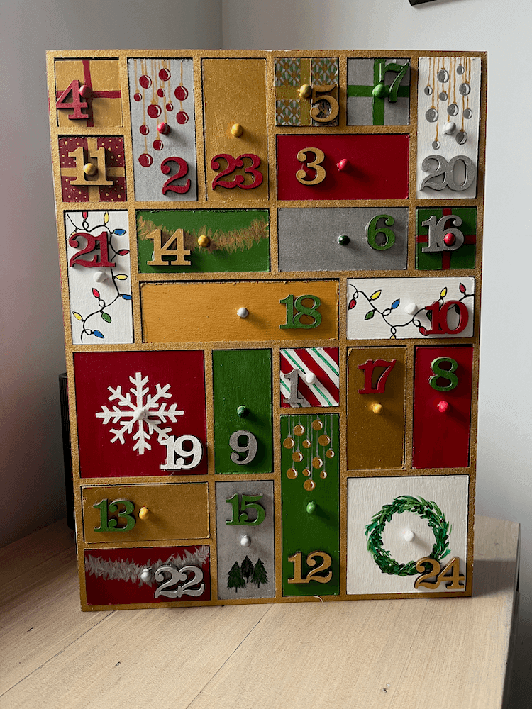 A wooden advent calendar with 24 various-sized drawers. The drawers have been painted red, gold, silver, green and white, and decorated with christmas motifs such as trees, tinsel, wreaths, candy cane stripes and the smallest drawers have been painted to look like presents.