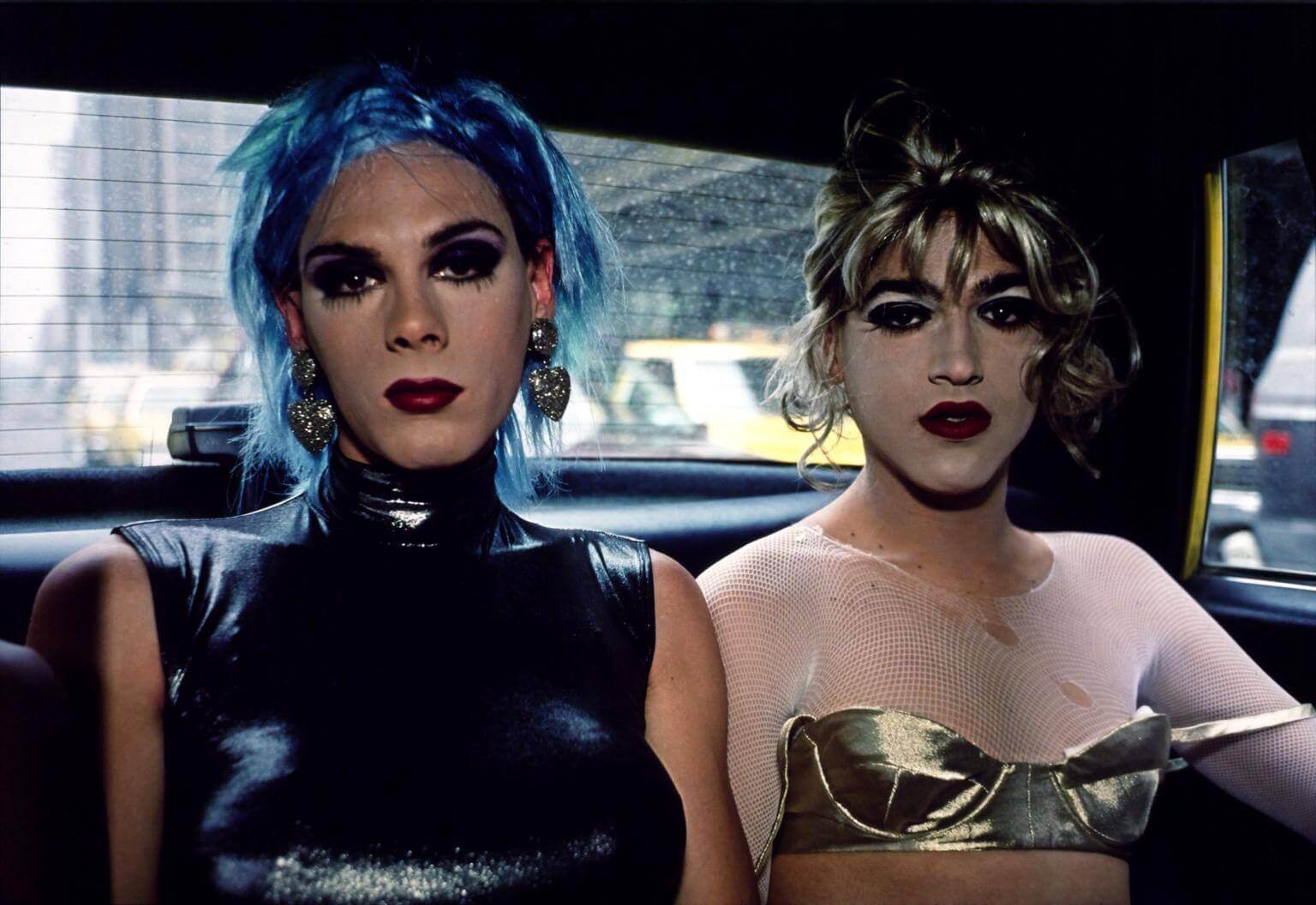 Two drag queens sit in the back of a taxi. One of them is wearing a tight metallic tank sleeveless top and has a short, choppy blue wig; she is wearing large silver heart-shaped earrings, thick dark eyeshadow and deep crimson lipstick. The second drag queen is wearing a long-sleeved ripped white fishnet crop top under a metallic bra, with a curly blonde updo wig, very pale face powder with dark eyeshadow and dark red lips. The straps of her bra are hanging off her shoulders.