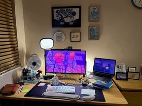 A photo of my desk in the evening. I have a monitor with a lo-fi style purple and blue illustration of a telegraph pole, a desk fan, and a space themed desk mat with a pastel coloured keyboard on it. THere is a mug with some different types of computers on it. I have an Elgato key light air behind my monitor and there's a leather-bound notebook on the desk. Behind the monitor are some pictures (an illustration of the indo-european and uralic language families as a tree, two risograph prints of illustrations of the Barbican conservatory, and a framed print of a caterpillar that says' I do not care, I will write whatever I want and post it online') and a framed cross-stitch sampler featuring the international phonetic alphabet. Next to the desk is a set of framed prints that are just propped up on a set of drawers.