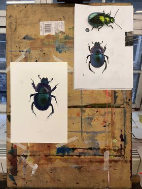 A photograph of a paint-covered board with a printed reference picture of two iridescent beetles clipped to it. The top beetle is a bright gold-green mix, and the bottom beetle is a purple and blue mix. Underneath the reference photo, a piece of paper is taped to the board with a painting of the bottom beetle. It is slightly messy, but the colours are accurate and the blending is quite good.