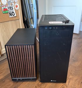 On the left, a small form factor black mesh computer case, with a slatted wood panel on the front. On the right, a much larger PC tower, coffee-splattered, with loose screws sitting on top. In the background, some stickers on a corkboard: one that says Perceivable & Operable & Understandable & Robust & Punch Nazis, a Keith Haring New York sticker, and one that says 'My heart says yes but my vocal range says no'.