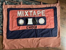 A peach-coloured fabric banner with a navy blue fabric cassette tape appliquéd on top. It's emblazoned with the words 'MIXTAPE CHOIR'. It is horribly wrinkled.