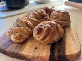 Small knotted cardamom buns sit carefully posed on a wooden board. They're glazed, and dusted with sugar.