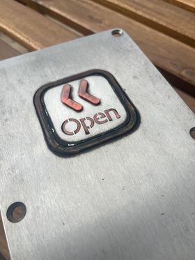 A very grimy metal plate with a button. The button is a square with rounded corners, the plastic ring around the button so dirty it looks black. The button itself has a pair of raised chevrons pointing left, and the word 'Open'.