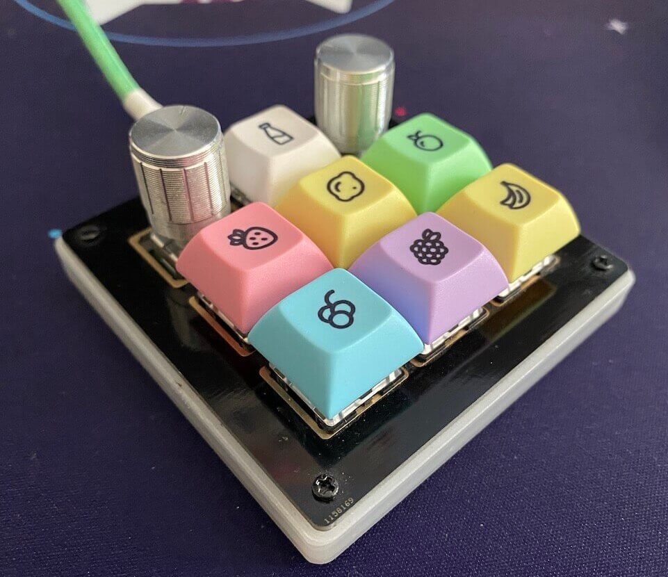 An angled view of the BDN9 macropad. It is a black square with two rotary knobs at the top, and 7 keys that are pastel coloured with fruit legends. There is a green cable plugged into it.