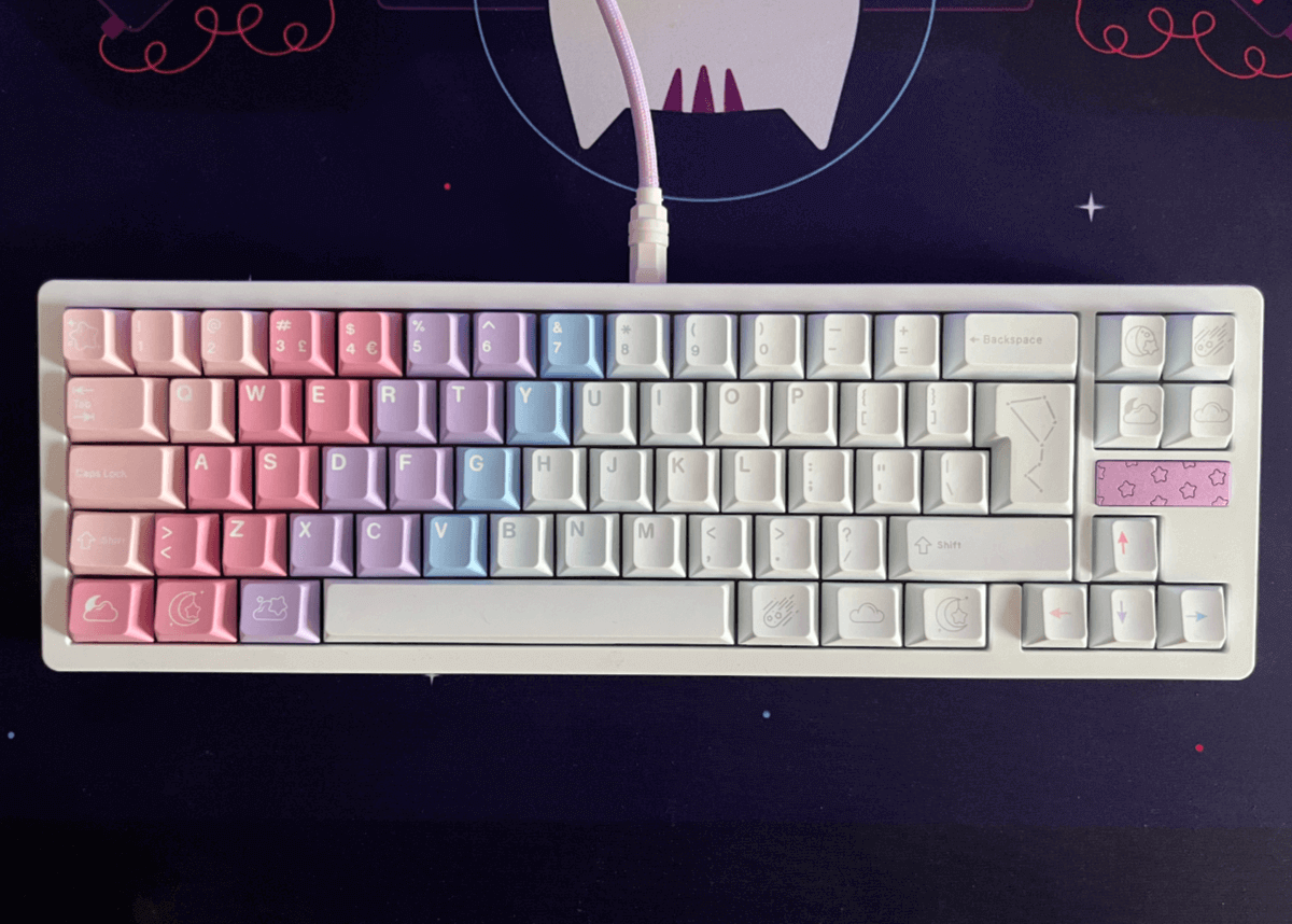 A top-down view of a mechanical keyboard with ISO layout. Its keycaps form a sort of gradient, colours in stripes going diagonally left to right from peach to pink to purple to blue to white. There is also a small purple plate on the keyboard with stars engraved on it. SOme of the keys have cute symbols on like stars, moons, clouds and meteors; the ISO enter key has a constellation on it. The keyboard sits on top of a deskmat with a cat in space on it.