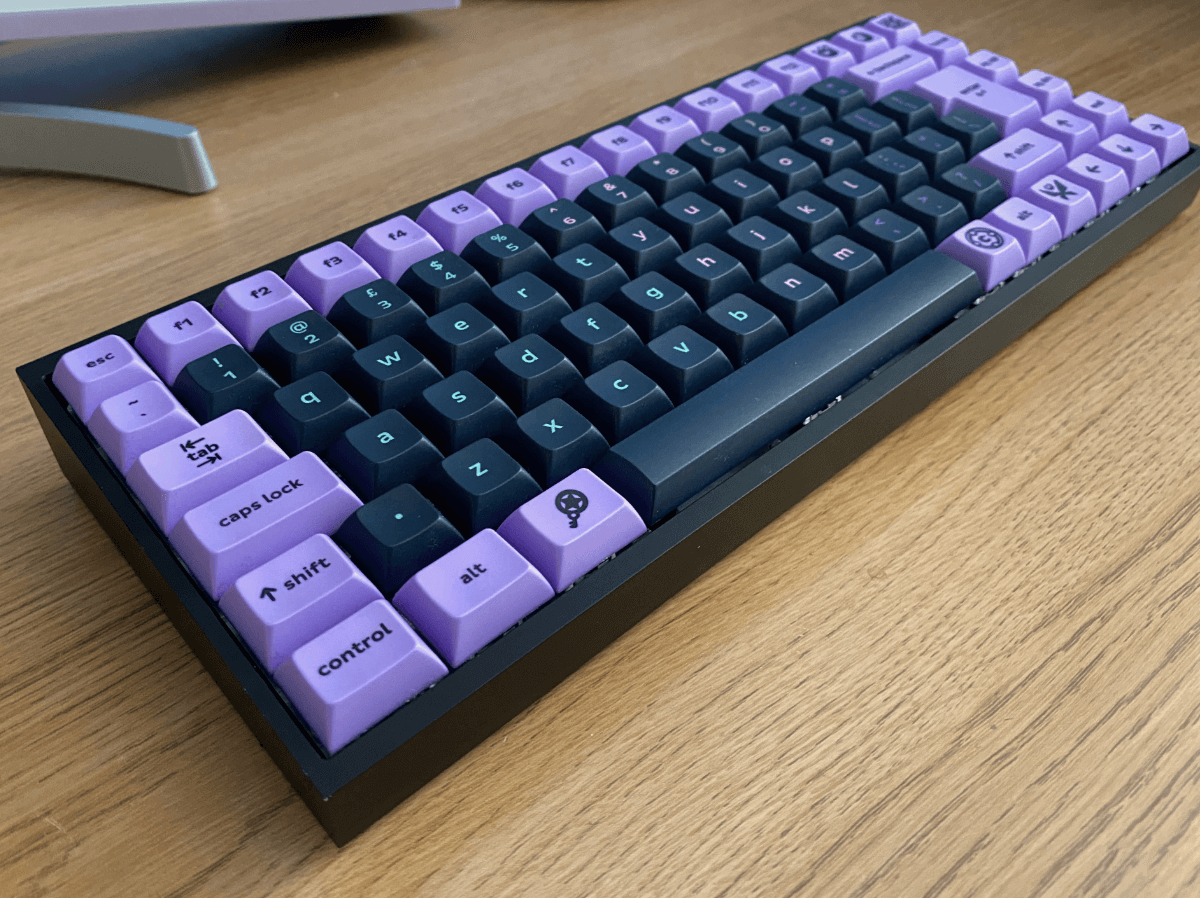 The KBDFans Tofu65 keyboard with black aluminium case. Its keycaps are purple and black, and the alpha key legends have a gradient from turquoise to peach. It is in Mac ISO layout with a split left shift.