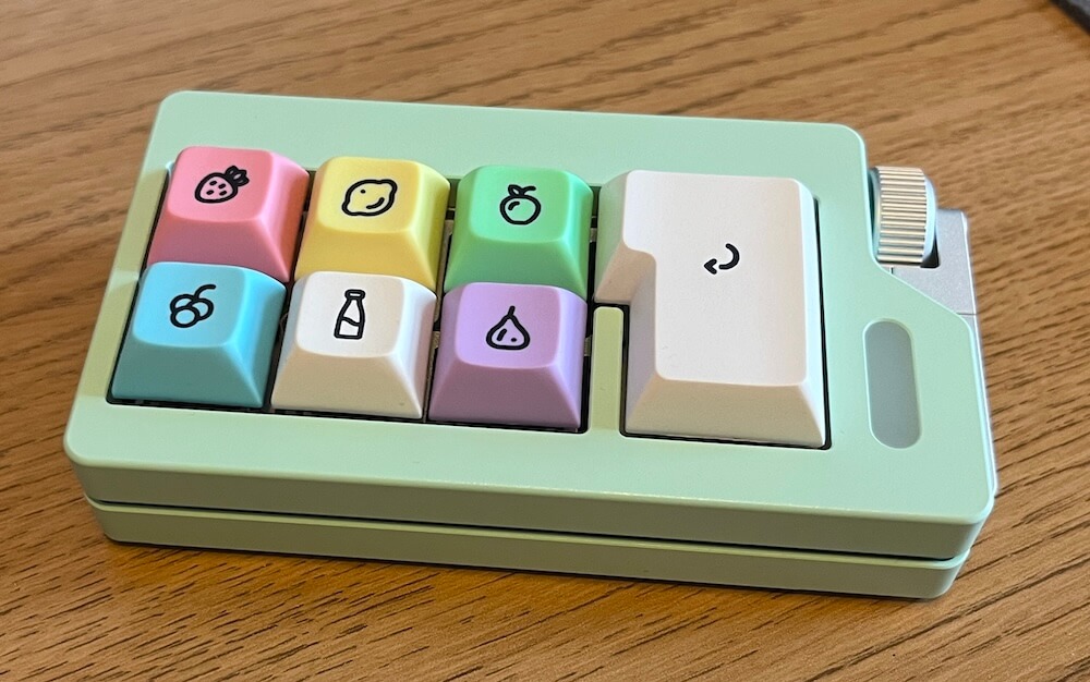 A mint green macropad with six regular sized keyboard keys and an ISO enter key, plus a silver knob on the side. The keys are decorated with fruits.