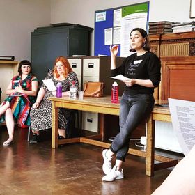 Sophie leans against a desk, hand in the air to indicate changes in pitch, mouth in an ooh shape as she teaches a song. There are two seated people visible and more out of shot.