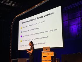 Emily Anhalt stands in front of a slide titled 'Emotional Fitness Survey Questions'. 'Do you like to be praised in public or in private? How do you like to receive feedback? How do you like to be cared for or cheered up during a tough time? How would I know if you were feeling overwhelmed? How do you like your birthday to be celebrated?'