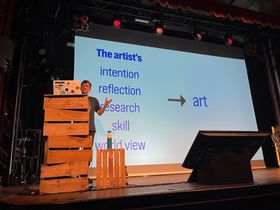 Hidde de Vries on stage in front of a slide that says 'The artist's intention, reflection, research, skill, world view' with a right arrow pointing to the word 'art'