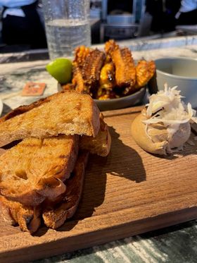 Golden slices of toast are stacked on a wooden board next to a scoop of caramel-coloured mushroom parfait. The parfait has shavings of white mushroom on top. In the background, out of focus, is a bowl of dark brown crispy corn ribs, served with a wedge of lime.